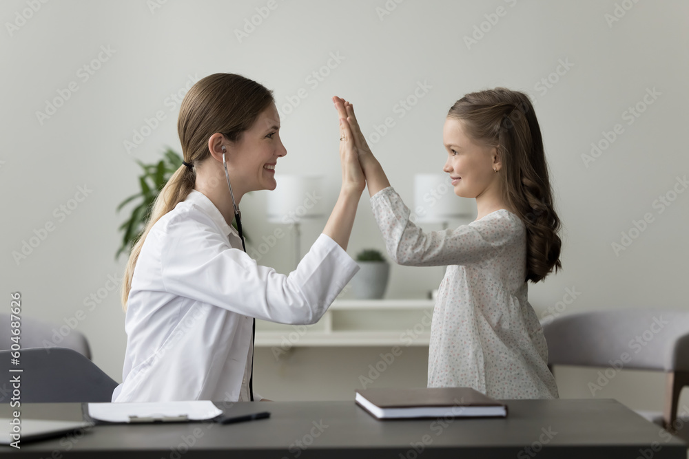 Friendly female pediatrician gives high five to little girl patient during visit in clinic. GP finish consultation with child, congratulates her with hand gesture, celebrate good results after therapy Friendly female pediatrician gives high five to little girl patient during visit in clinic. GP finish consultation with child, congratulates her with hand gesture, celebrate good results after therapy
