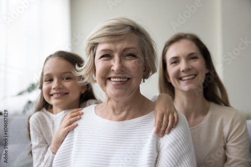 Attractive optimistic mature 60s woman looks at camera pose for picture with lovely granddaughter and young adult daughter. Caucasian female relatives feel happy, portrait of intergenerational family