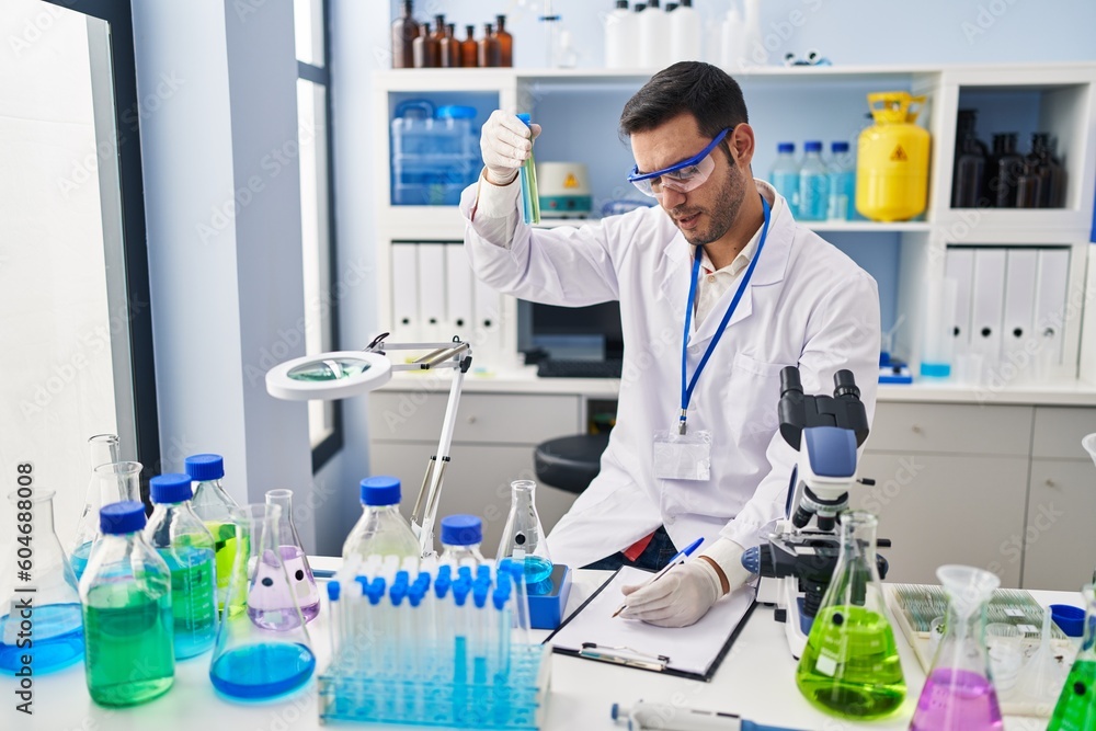 Fototapeta premium Young hispanic man scientist holding test tubes writing report at laboratory