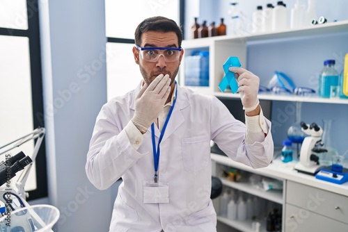 Young hispanic man with beard working at scientist laboratory holding blue ribbon covering mouth with hand  shocked and afraid for mistake. surprised expression