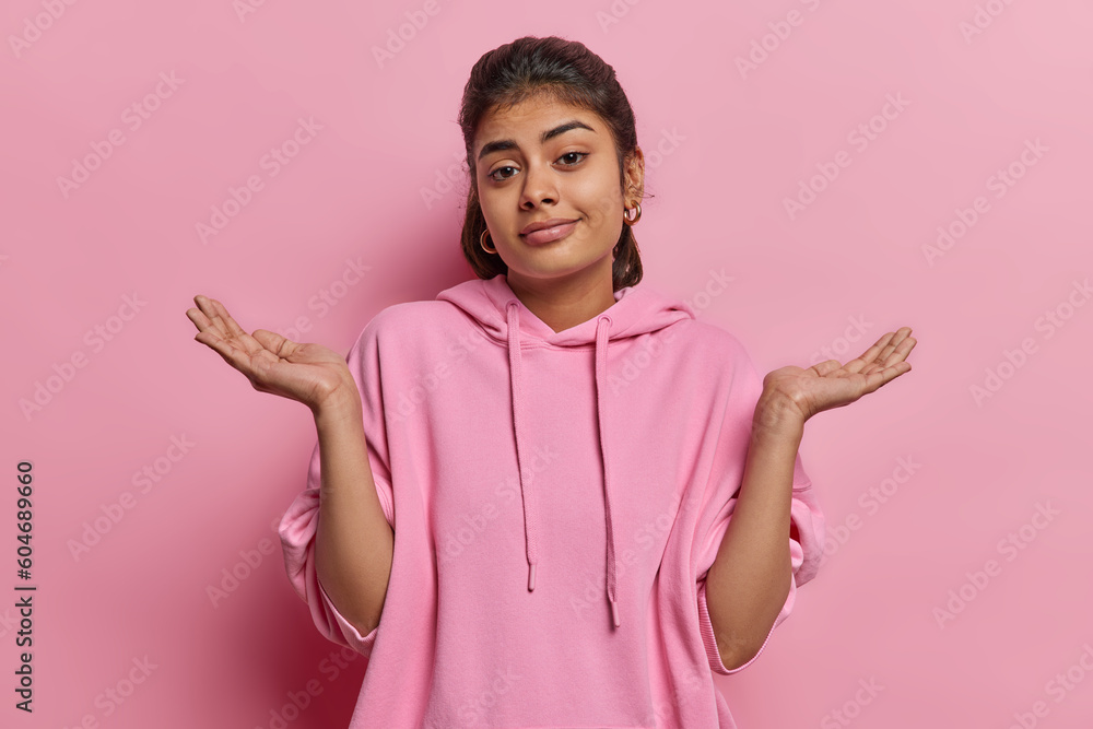 Clueless indecisive Indian woman spreads palm shrugs shoulders looks clueless at camera dressed in casual sweatshirt cannot decide or answer your question isolated over pink background faces dilemma - obrazy, fototapety, plakaty 