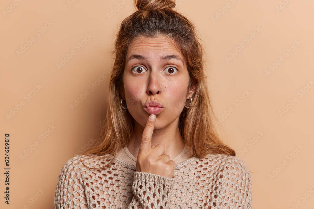 Lovely speechless woman has widely opened eyes has lips folded concentrated at camera with wondered expression wears casual knitted jumper isolated over brown background. Human facial expressions Lovely speechless woman has widely opened eyes has lips folded concentrated at camera with wondered expression wears casual knitted jumper isolated over brown background. Human facial expressions