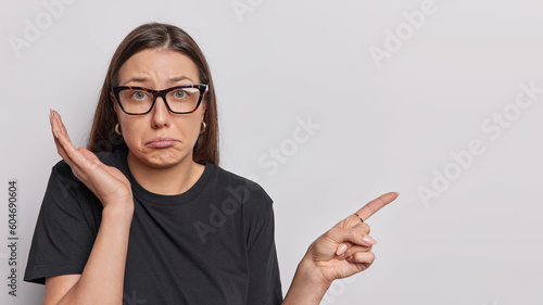 Horizontal shot of frustrated woman purses lips keeps palm raised points index finger aside on blank space tells bad news shows mock up space for your advertisement wears spectacles and black t shirt