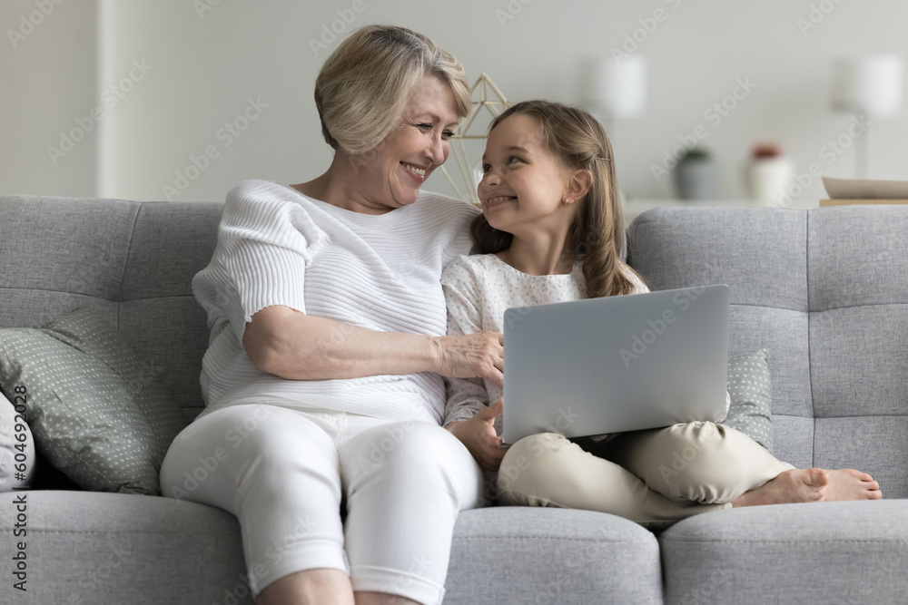 Multi generational family, grandma and granddaughter sit on couch with wireless computer, look at each other with tenderness and love, enjoy leisure on internet and time together, watch on-line movies Multi generational family, grandma and granddaughter sit on couch with wireless computer, look at each other with tenderness and love, enjoy leisure on internet and time together, watch on-line movies