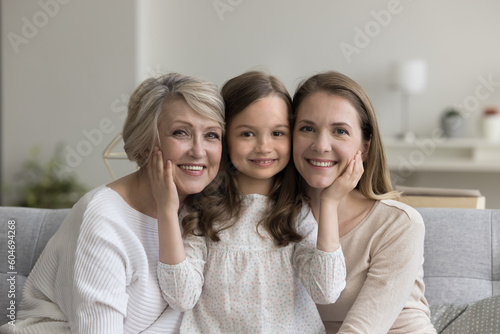 Portrait of happy cute little 6s girl posing for camera with retired elderly granny and young attractive mother sit together on couch smile looking at camera, showing care, feeling unconditional love