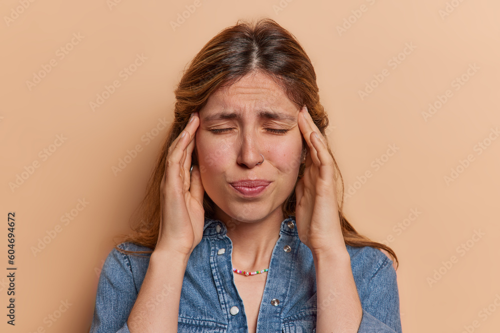 Frustrated young European woman suffers from headache keeps hands on temples to reveal pain got things messed up tries to calm down during panic wears denim shirt isolated over brown background Frustrated young European woman suffers from headache keeps hands on temples to reveal pain got things messed up tries to calm down during panic wears denim shirt isolated over brown background