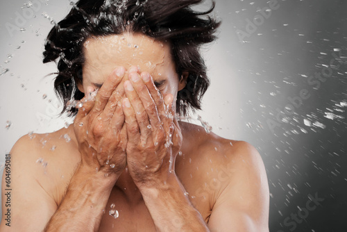 Unrecognizable shirtless man washing face with water splashing around against grey background