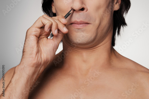 Close-up of young shirtless man plucking hair from nose and grimacing against grey background