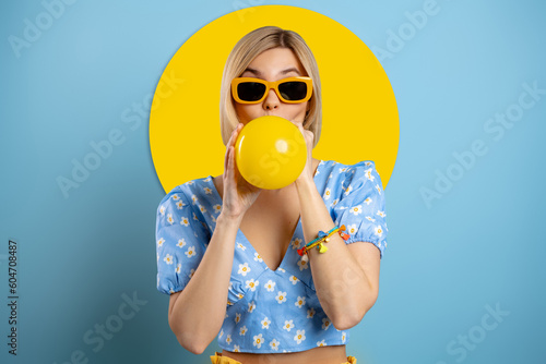 Playful young woman blowing up a balloon while standing against blue background