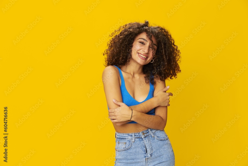 Woman with curly afro hair in a blue t-shirt on. yellow background signs with her hands, look into the camera, smile with teeth and happiness, copy space Woman with curly afro hair in a blue t-shirt on. yellow background signs with her hands, look into the camera, smile with teeth and happiness, copy space