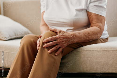 Elderly woman severe pain in her leg sitting on the couch, health problems in old age, poor quality of life. Grandmother with gray hair holds on to her sore knee, problems with joints and ligaments.