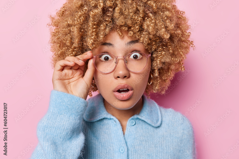 Portrait of shocked emotional curly haired woman stares through spectacles gazes with stupefaction hears surprising news dressed in blue sweater isolated over pink background. Human reactions concept - obrazy, fototapety, plakaty 