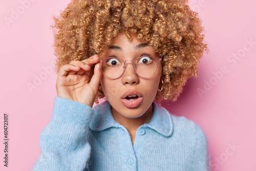 Portrait of shocked emotional curly haired woman stares through spectacles gazes with stupefaction hears surprising news dressed in blue sweater isolated over pink background. Human reactions concept