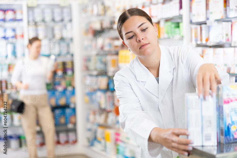 Young woman pharmacist in uniform arranges products on shelves in pharmacy