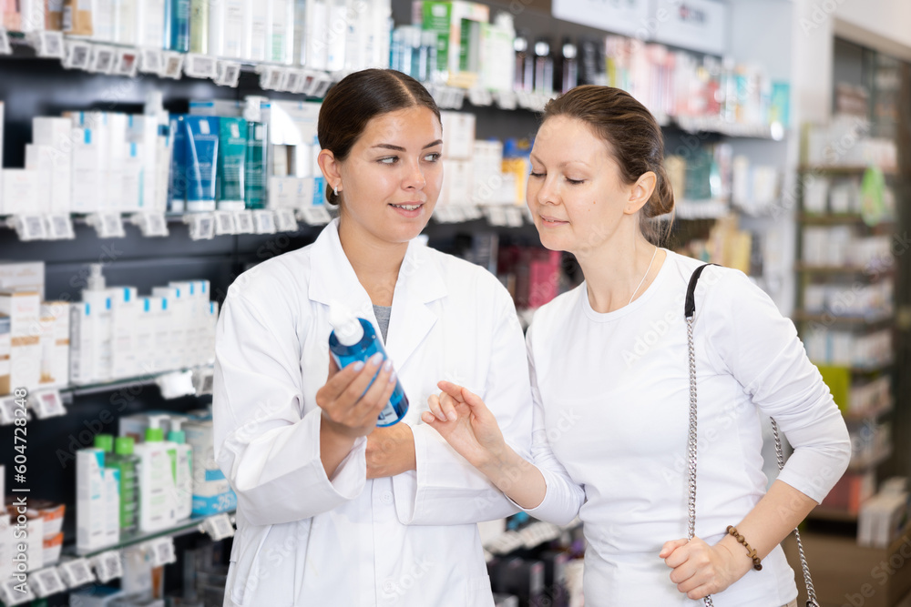 Female pharmacist in uniform helping female shopper in casual wear choose cosmetic product in pharmacy