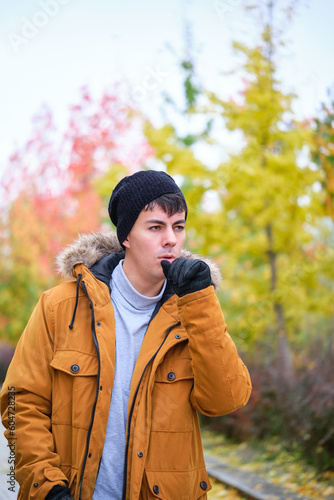 Young man in coat, hat and gloves coughing on a cold autumn day at street. © Ladanifer
