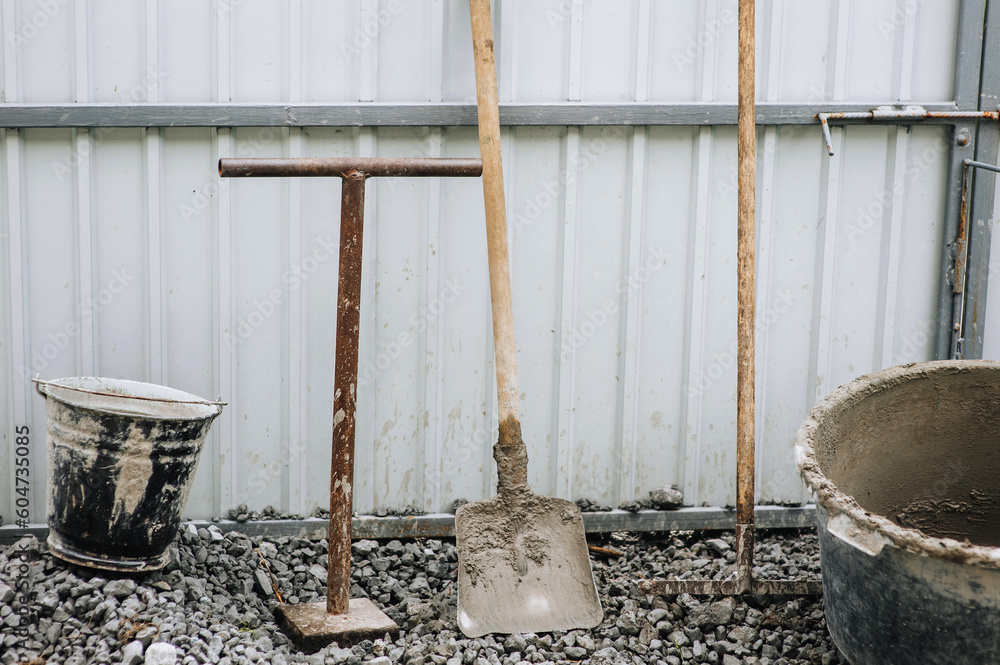 Construction tools: a bucket, a shovel, a rake, a concrete mixer in concrete, cement stand on stones against the background of a house wall. Close-up photo. Construction tools: a bucket, a shovel, a rake, a concrete mixer in concrete, cement stand on stones against the background of a house wall. Close-up photo.