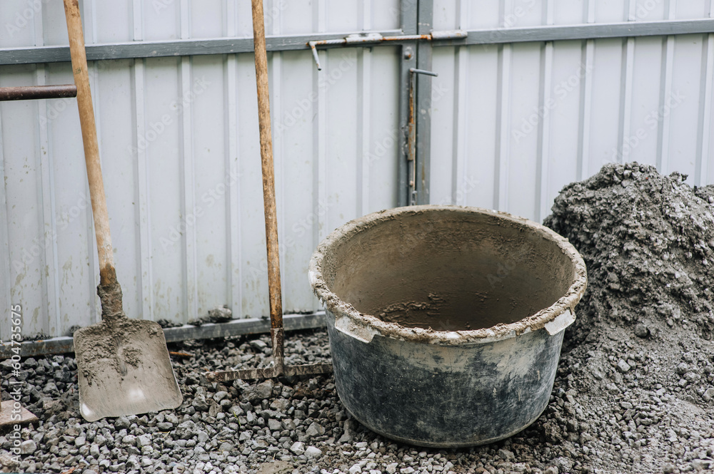 Construction tools: a bucket, a shovel, a rake, a concrete mixer in concrete, cement stand on stones against the background of a house wall outdoors. Construction tools: a bucket, a shovel, a rake, a concrete mixer in concrete, cement stand on stones against the background of a house wall outdoors.
