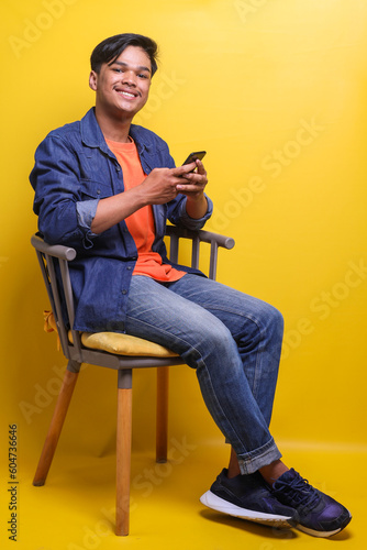 Asian cheerful young man using smart phone, sit on chair and looking at camera smiling isolated on yellow background
