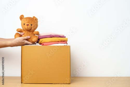 Kid Donation, Charity, Volunteer, Giving and Delivery Concept. Hand donate Bear doll and Clothes into cardboard box at home for support and help poor, refugee and homeless people. Copy space for text © Jo Panuwat D