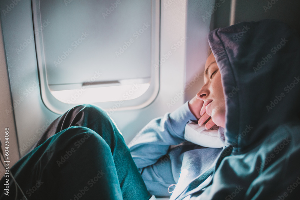 Tired blonde casual caucasian woman wearing sporty hoodie napping on seat while traveling by airplane. Commercial transportation by planes. Authentic image of real people - obrazy, fototapety, plakaty 