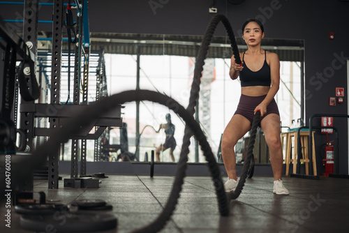 Strong Asian woman doing exercise with battle rope at cross-fit gym. Athlete female wearing sportswear workout on grey gym background with weight and dumbbell equipment. Healthy lifestyle.