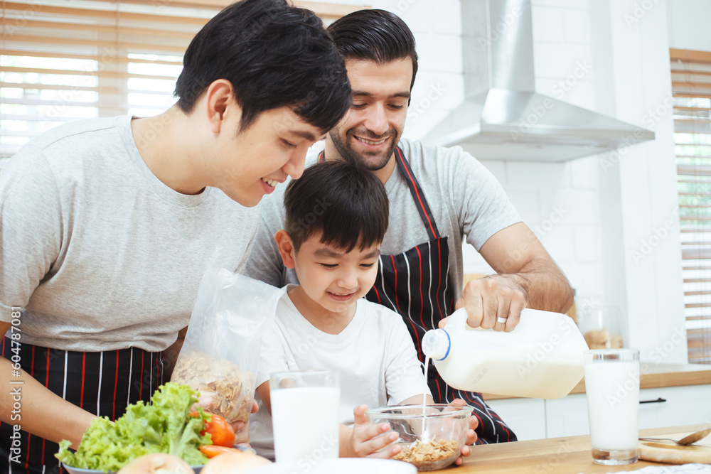 Happy cheerful gay couple with their adopted little son enjoy making a corn flakes with fresh milk for breakfast in the kitchen together. Happy LGBTQ+ family with adopted child concept.