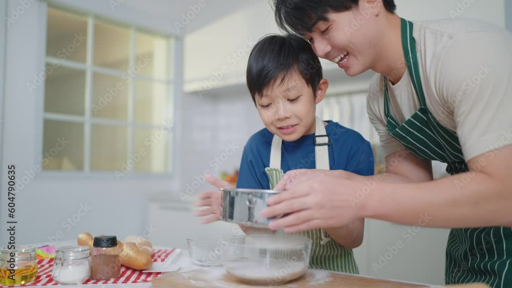 Happy asian father teaching little son sifts flour through sieve in glass bowl for cooking or baking enjoying weekend in kitchen room at home together. Family time concept