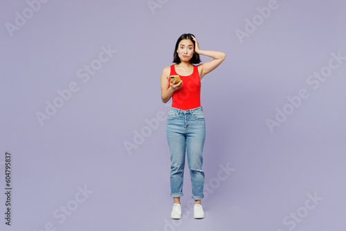 Full body scared sad young woman of Asian ethnicity she wear casual clothes red tank shirt hold head use mobile cell phone isolated on plain pastel light purple background studio. Lifestyle concept.