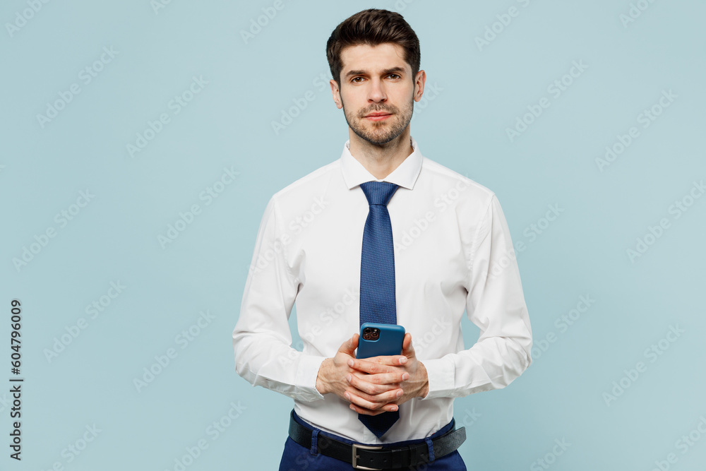 Young confident employee business man corporate lawyer wear classic formal shirt tie work in office hold in hand use mobile cell phone isolated on plain pastel light blue background studio portrait. Young confident employee business man corporate lawyer wear classic formal shirt tie work in office hold in hand use mobile cell phone isolated on plain pastel light blue background studio portrait.