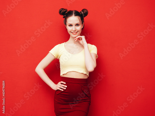 Young beautiful smiling female in trendy summer yellow t-shirt and skirt. Carefree woman with two horns hairstyle posing near red wall in studio. Positive model having fun