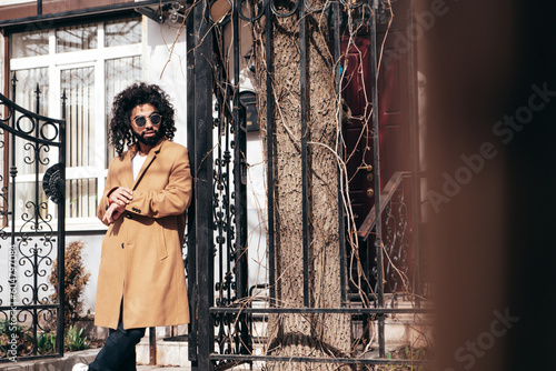 Handsome smiling hipster model. Unshaven Arabian man dressed in brown coat clothes. Fashion male with long curly hairstyle posing outdoors in street. Cheerful and happy. In sunglasses. At sunny day