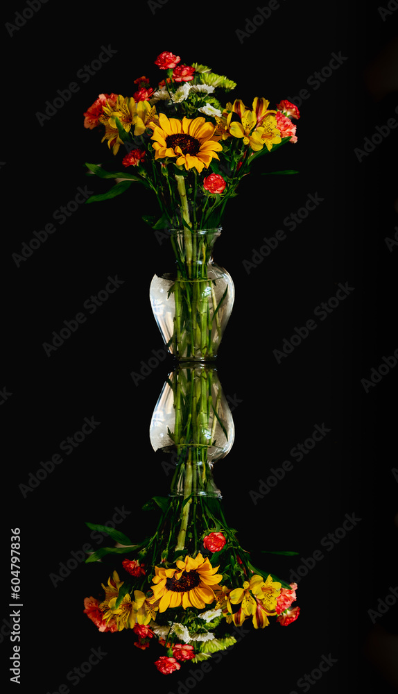 Obraz premium Beautiful view of vibrant bouquet of red carnations, sunflowers and chrysanthemum in glass vase reflecting in glass; black background