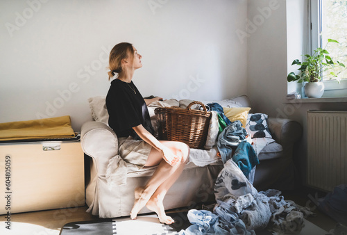 Thoughtful woman sitting on sofa with cluttered laundry at home