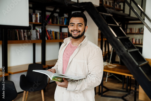 Cheerful indian man holding textbook while standing in library photo