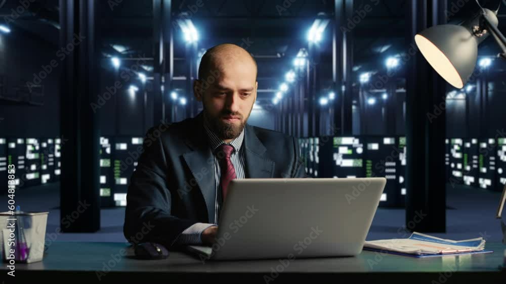 IT engineer in suit working on laptop, sitting at desk in modern server room with cloud computing hardware. Businessman in data center with networking and security information. 3d render animation. IT engineer in suit working on laptop, sitting at desk in modern server room with cloud computing hardware. Businessman in data center with networking and security information. 3d render animation.