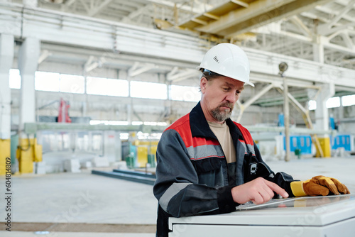 Engineer in helmet touching the screen of industrial computer and controlling the process of production in workshop