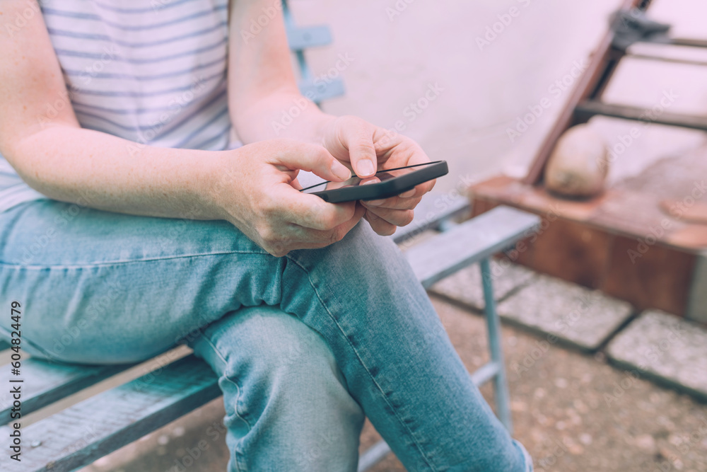 Casual female person wearing jeans trousers using smartphone device outdoors while sitting on the bench in house back yard