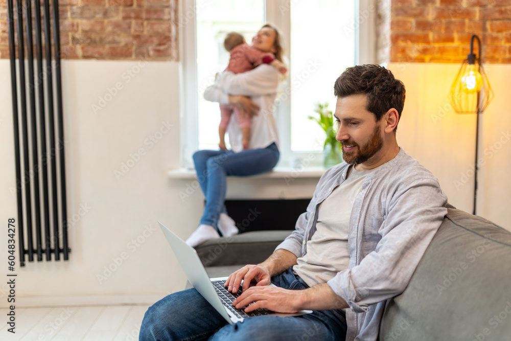 Bearded man working on computer, wife and toddler daughter on the background hugging. Bearded man working on computer, wife and toddler daughter on the background hugging.