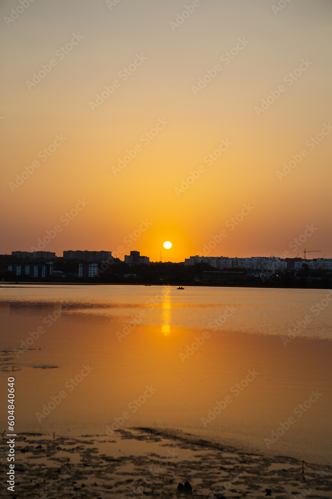 Far away, against the backdrop of a shui town, a fisherman is fishing on the lake from an inflatable small boat, against the backdrop of a beautiful orange sunset.
