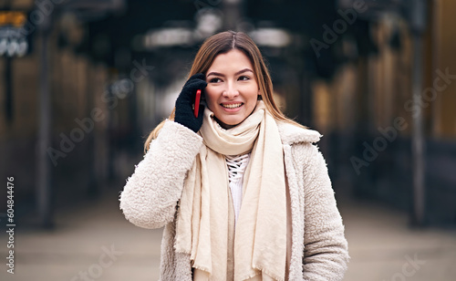 Smiling millennial student woman walking on city street and discussing the daily topics in a phone call with a tram stop in the background - Teenager girl using telephone outside in jacket