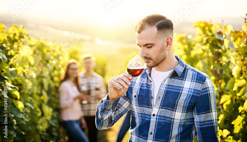 Portrait of a young, millennial vintner holding and smelling a glass of organic bio red wine outdoors in a vineyard with his friends in the background - Vine-growing, and wine-tasting concept © napeter