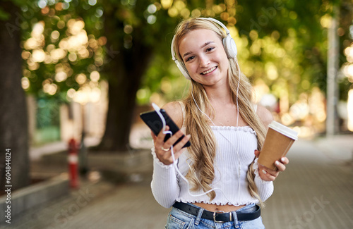 Cheerful teenager student girl walking on a street with a coffee to go paper cup in her hand and listening to music with headphones from a smart phone