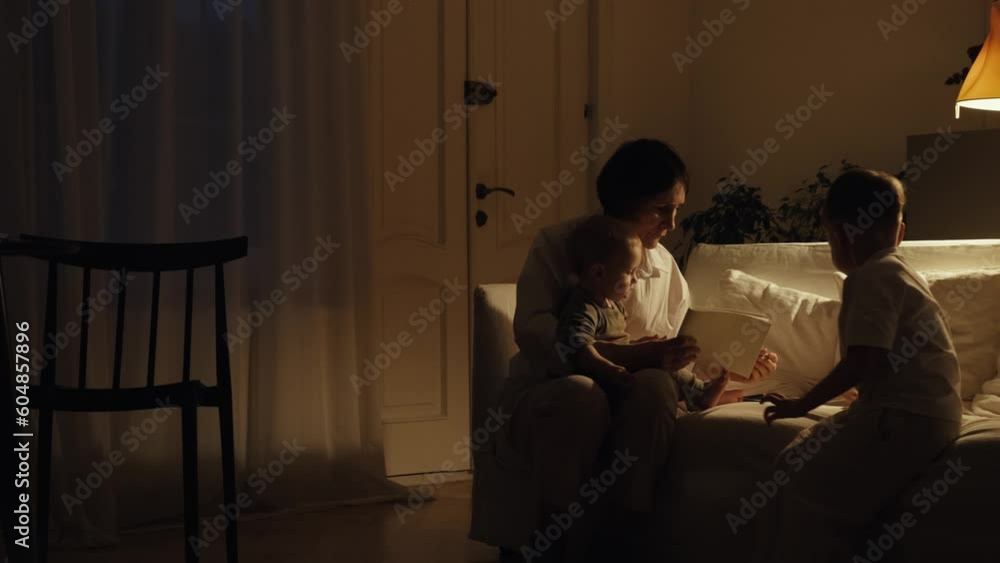Grandmother and two grandchildren are sitting on a white sofa and drawing with colored pencils. The little boy is sitting in his grandmother's arms. Family evenings. Grandmother and two grandchildren are sitting on a white sofa and drawing with colored pencils. The little boy is sitting in his grandmother's arms. Family evenings.