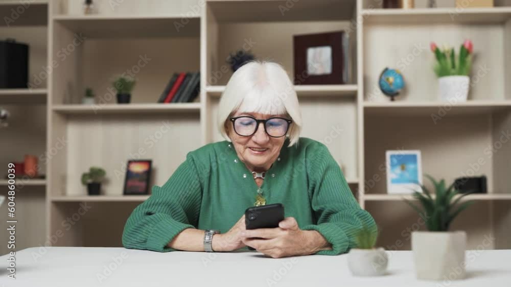 There is a wonderful elderly woman with short hair and glasses sitting at the table. She is holding a phone in her hands and enjoying what she is doing on it. Her face reflects positive emotions. There is a wonderful elderly woman with short hair and glasses sitting at the table. She is holding a phone in her hands and enjoying what she is doing on it. Her face reflects positive emotions.