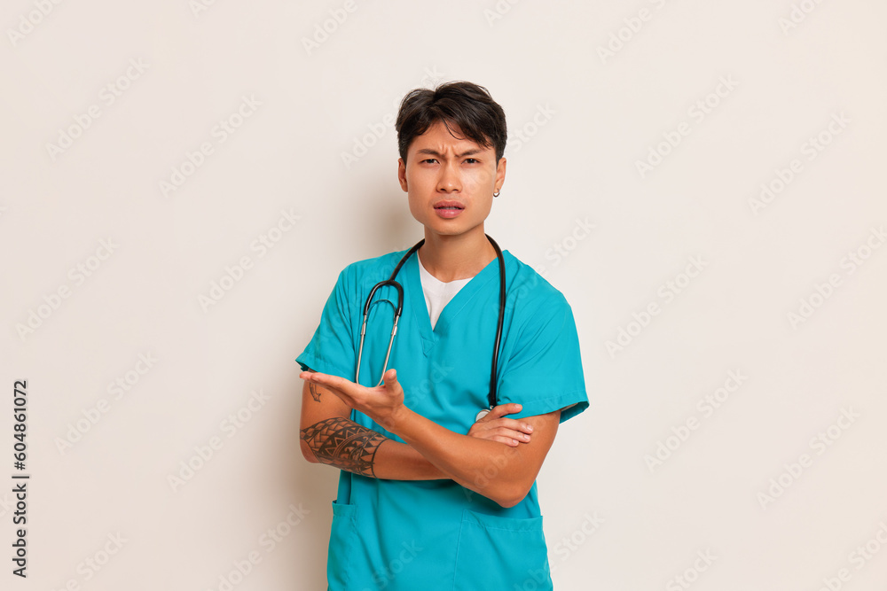  Serious young man in turquoise medical uniform stands with his arms folded on his chest with bewilderment on his face, medical care concept, copy space