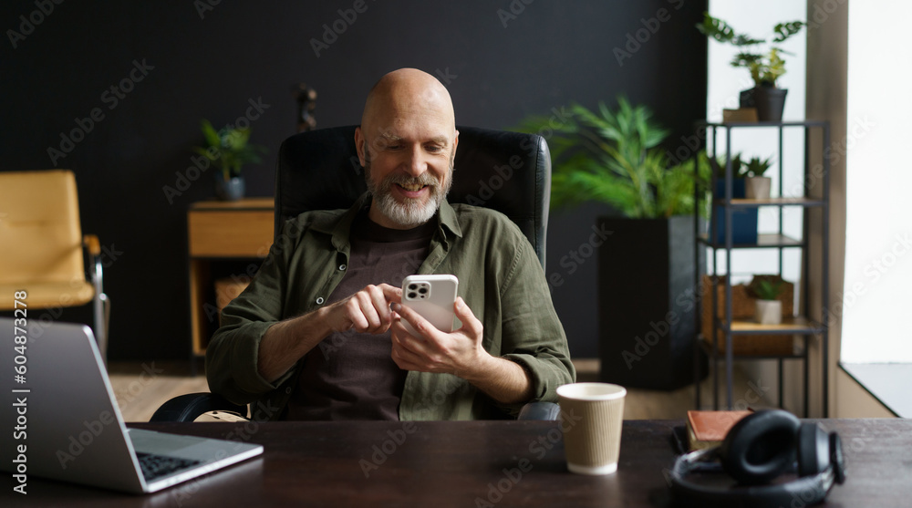 Bald and respectable man with gray beard. He captured in moment of joy, smiles while reading messages on phone. Computer, cup of coffee, and audio headphones are present on the table. Bald and respectable man with gray beard. He captured in moment of joy, smiles while reading messages on phone. Computer, cup of coffee, and audio headphones are present on the table.