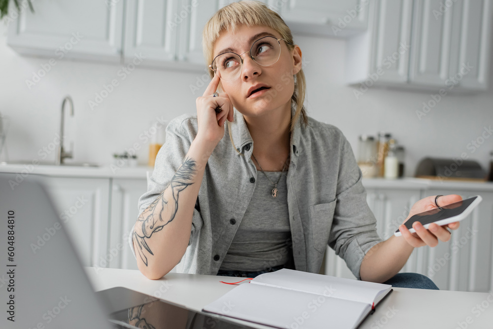pensive young woman with tattoo on hand and bangs holding smartphone with blank screen and pen near notebook and laptop on white table, blurred foreground, work from home pensive young woman with tattoo on hand and bangs holding smartphone with blank screen and pen near notebook and laptop on white table, blurred foreground, work from home
