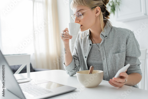 young woman with tattoo on hand and bangs holding smartphone and using laptop  drinking coffee while holding cup near and bowl wth cornflakes on table in modern kitchen  freelancer