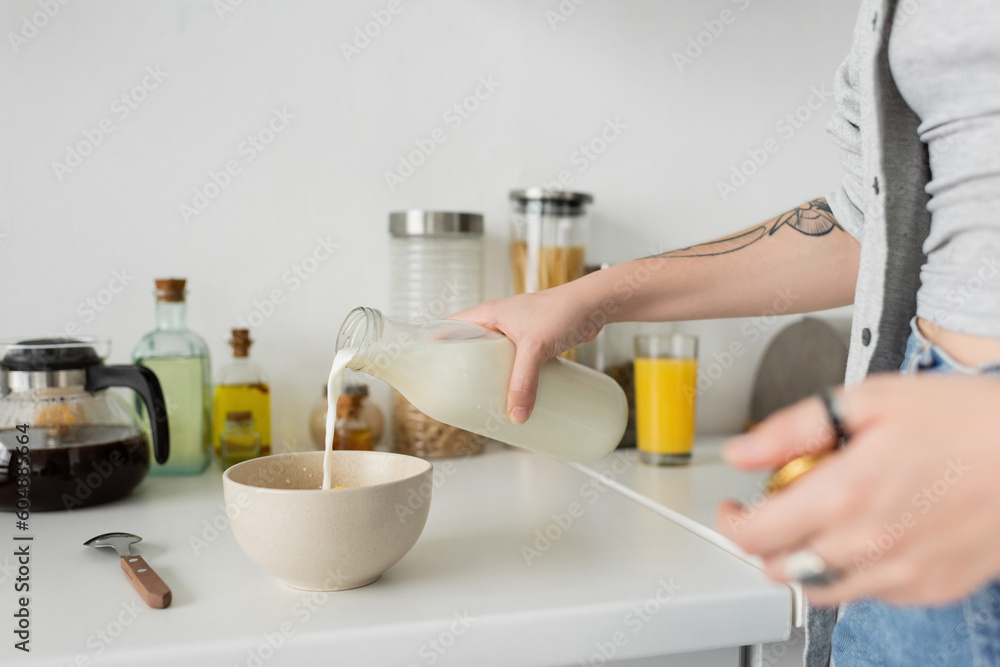 cropped view of tattooed young woman holding bottle while pouring fresh milk into bowl with cornflakes on kitchen worktop while making breakfast and standing in casual clothes in modern kitchen